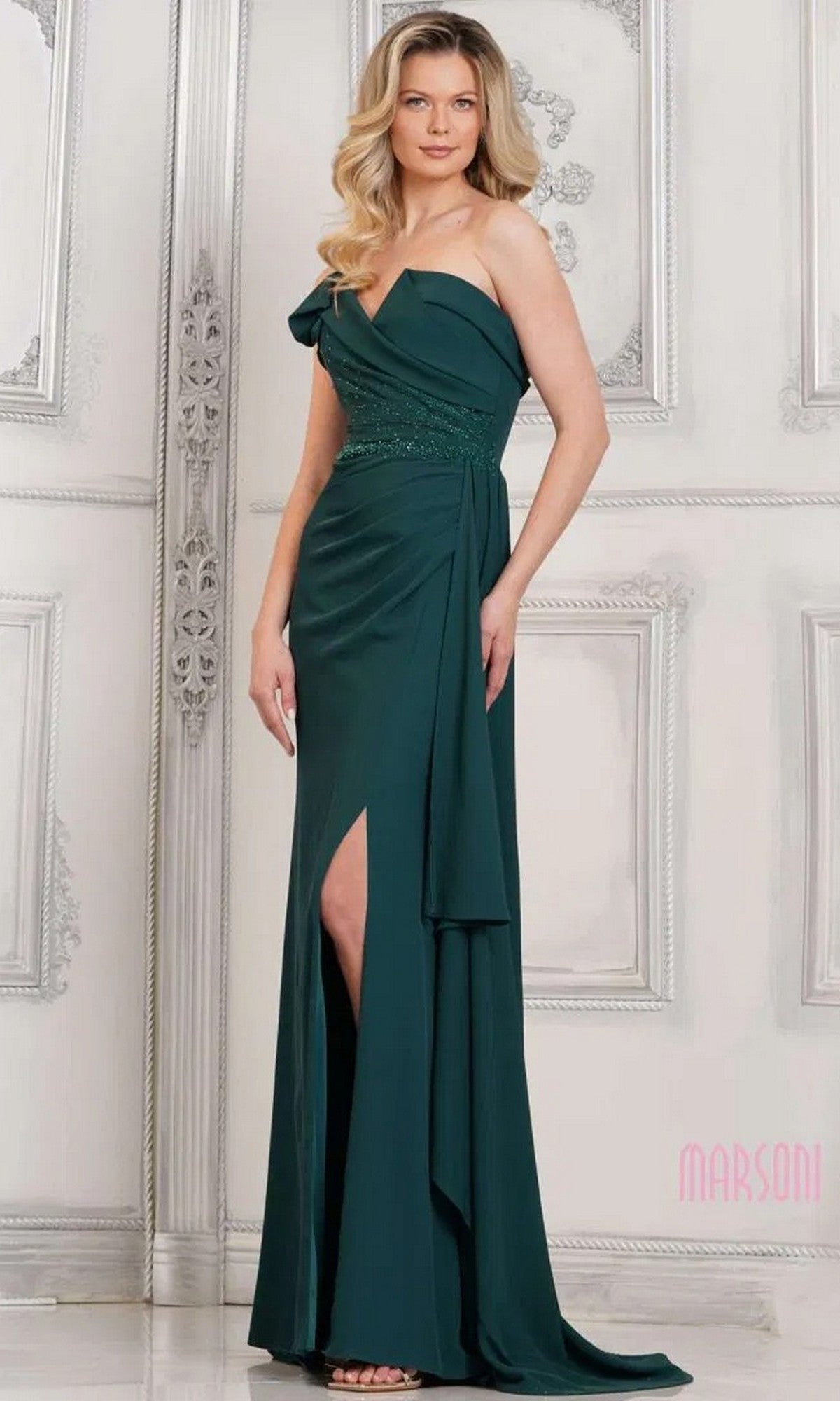A woman models the Marsoni MV1304 long formal dress in deep green, a floor-length, one-shoulder gown with a thigh-high slit, posing against an ornate white wall.
